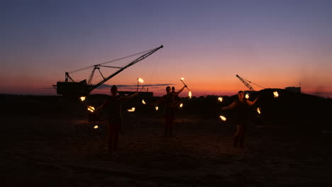 Eine-Gruppe-Von-Männern-Und-Frauen-Zeigt-Nachts-Eine-Feuershow-Auf-Dem-Sand-Vor-Dem-Hintergrund-Von-Feuer-Und-Turmdrehkränen.